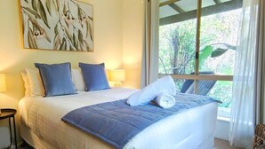 Una cama bien hecha con una manta y almohadas azules, una lámpara de noche y una ventana con vista a la vegetación.