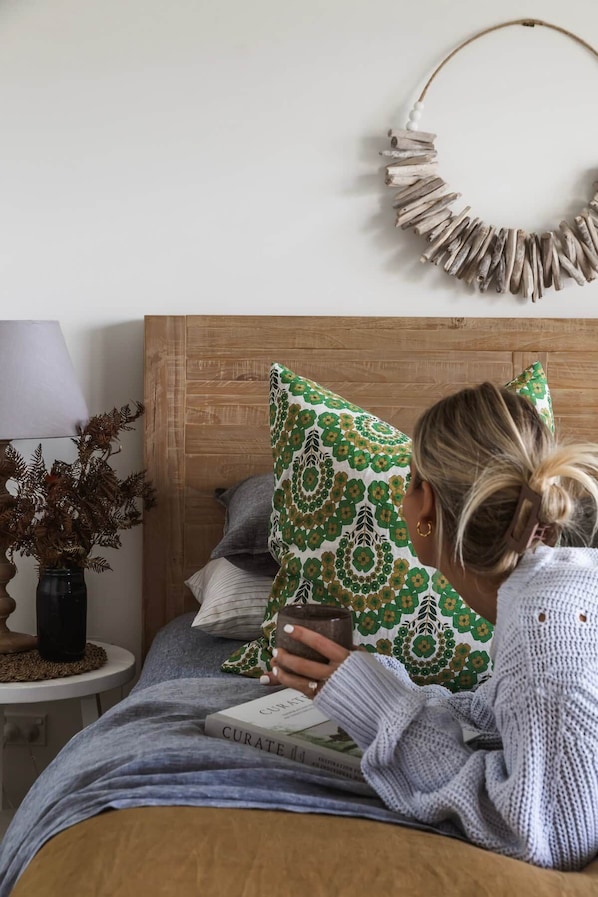 Una persona leyendo un libro en una cama con cabecera de madera, un adorno en la pared y una mesita con una lámpara y un jarrón de plantas secas.