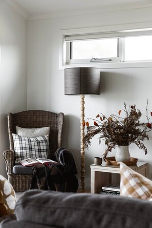 A corner room with a wicker chair, a floor lamp, a small table with a vase of dried branches, and a window above.
