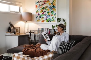 A person sitting on a couch reading a book in a room with a bookshelf, a lamp, and a painting on the wall.