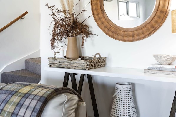 A staircase with a white wall, a round mirror, a vase with dried plants, a wicker basket, and a white trash bin.