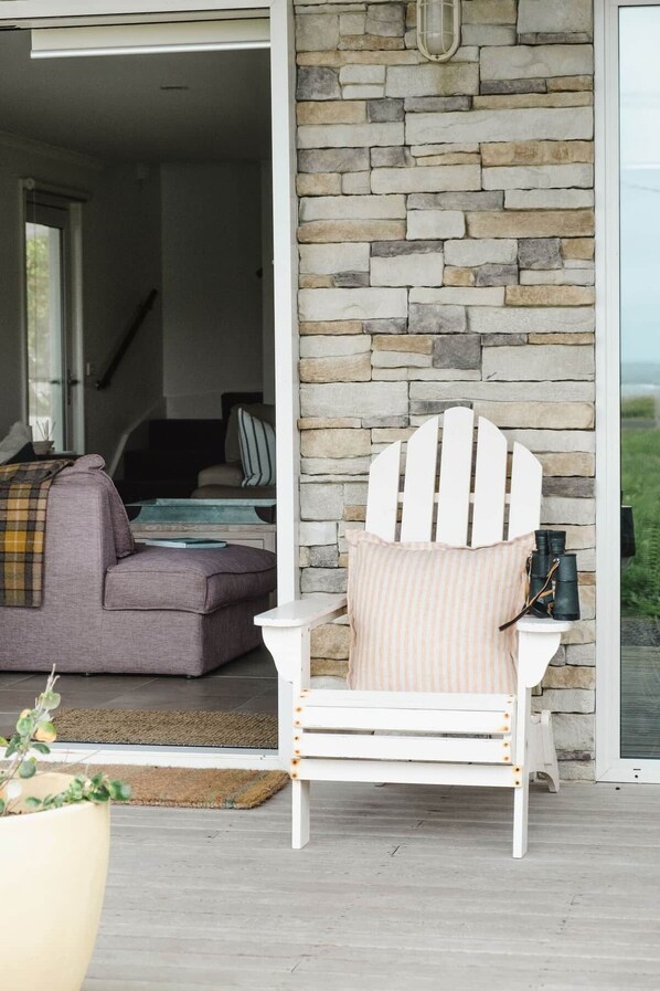 A white Adirondack chair with a striped cushion, positioned in front of a stone wall, with a view of a house interior in the background.