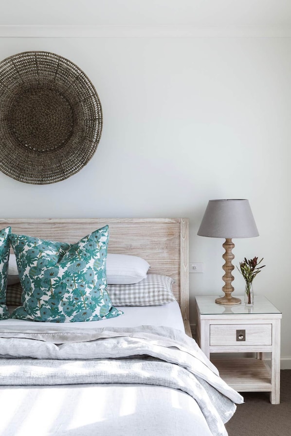 A neatly made bed with a checkered pillow and floral pillows, a bedside lamp, and a woven wall hanging.