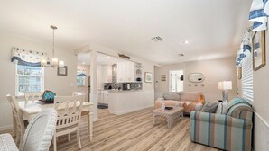 A spacious living area with a dining table, chairs, a sofa, and a coffee table. A kitchen with white cabinets and a stone backsplash is visible in the background.