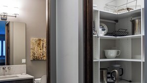 Ein modernes Badezimmer mit Holzboden, einem weißen Waschbecken und einer Toilette. Es gibt eine Regaleinheit mit Geschirr und einem Staubsauger.
