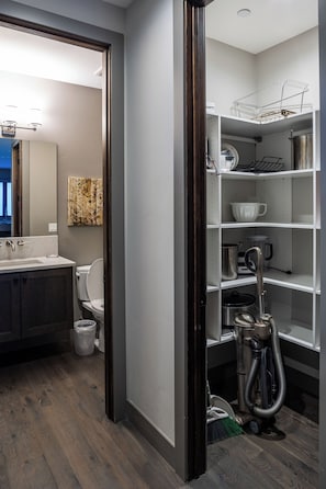 Un baño moderno con piso de madera, un lavamanos blanco y un inodoro. Hay una estantería con utensilios de cocina y un aspirador.