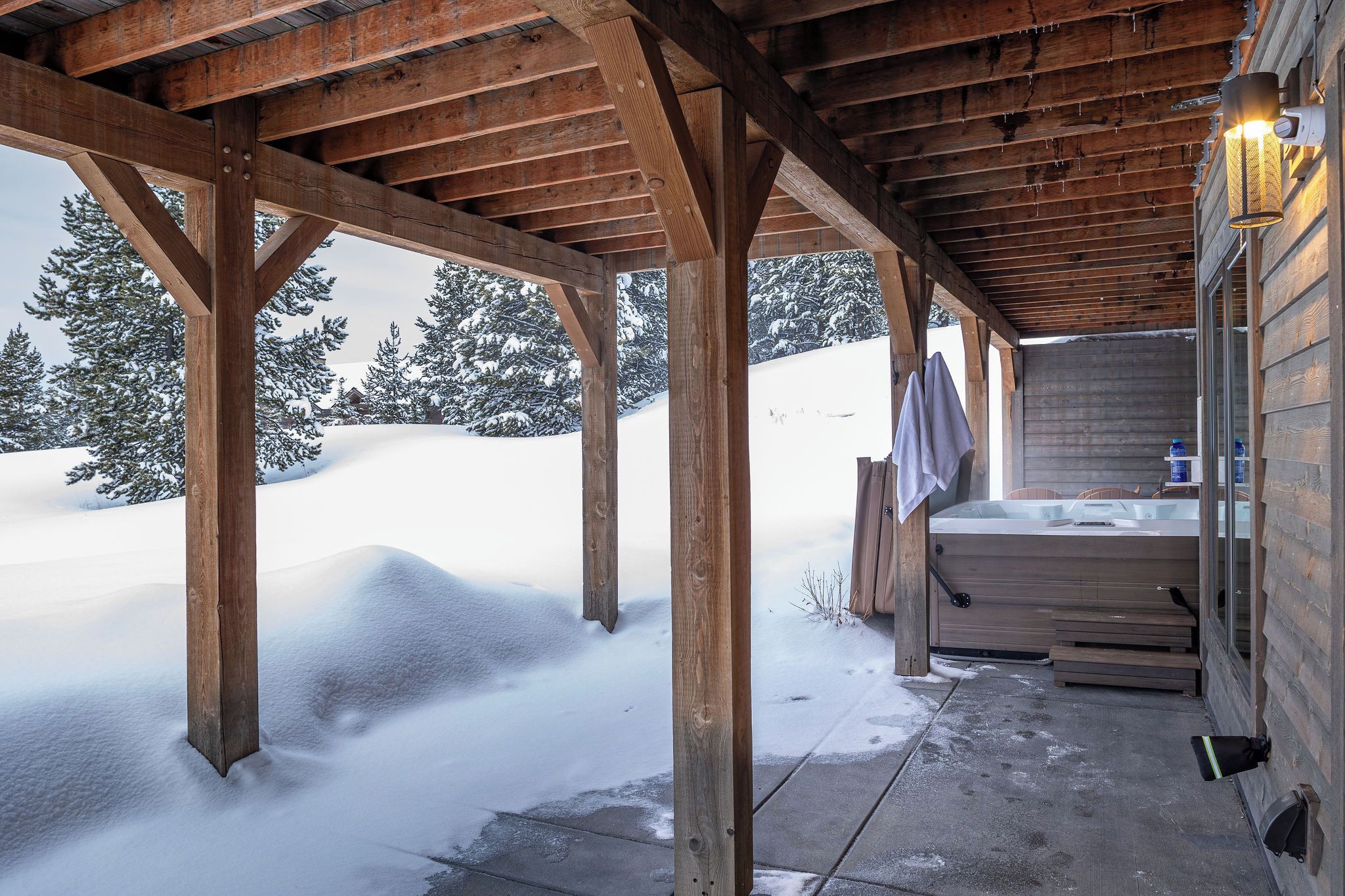 A wooden cabin with a covered porch, a hot tub, and a snowy landscape in the background.