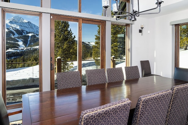 Un comedor con mesa de madera y sillas estampadas, ventanas amplias con vista a la montaña y una lámpara de araña moderna.