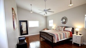 A spacious bedroom with a large bed, a ceiling fan, a bedside table with a lamp, and a mirror on the wall.