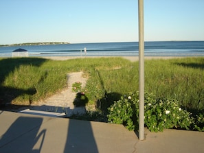 A concrete pathway leading to a beach with a grassy area and a metal pole. - Panoramic Views & Crashing Waves (Old Orchard Beach)