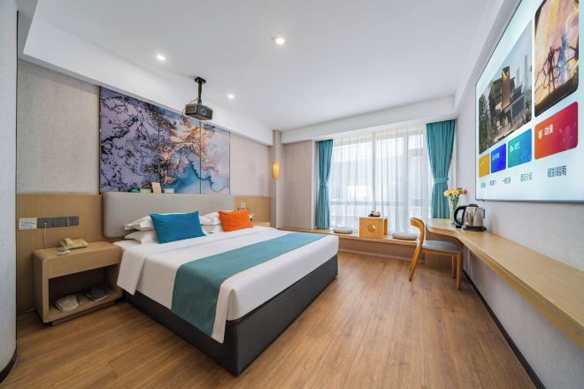 Quarto de hotel com cama, escrivaninha e cadeira, um quadro abstrato grande e uma janela com cortinas.