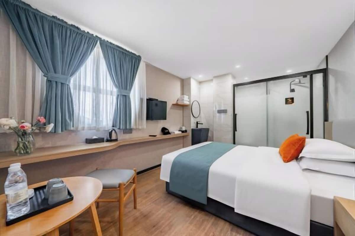 A hotel room with a bed, a desk, a chair, a TV, a mirror, and a shower area.