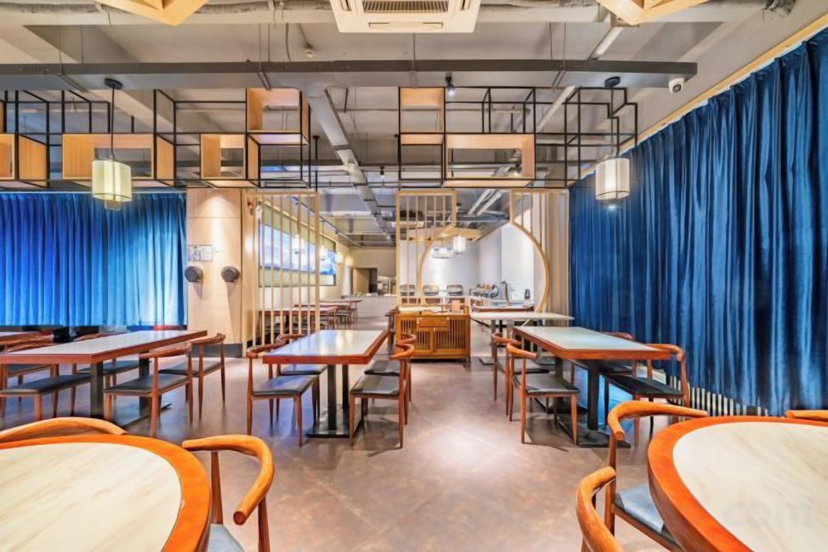Un interior de restaurante con varias mesas y sillas, una zona de cocina al fondo y cortinas azules.