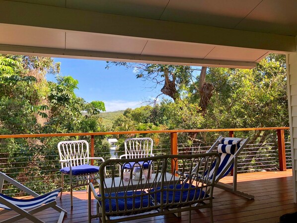 Un espace extérieur aménagé, abrité et doté de chaises et d’une table en métal, entouré d’une terrasse en bois et d’une rambarde métallique, offrant une vue sur un paysage verdoyant et luxuriant.