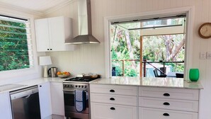 Una cocina compacta con muebles blancos, estufa a gas, lavavajillas y ventana con vista a un jardín.
