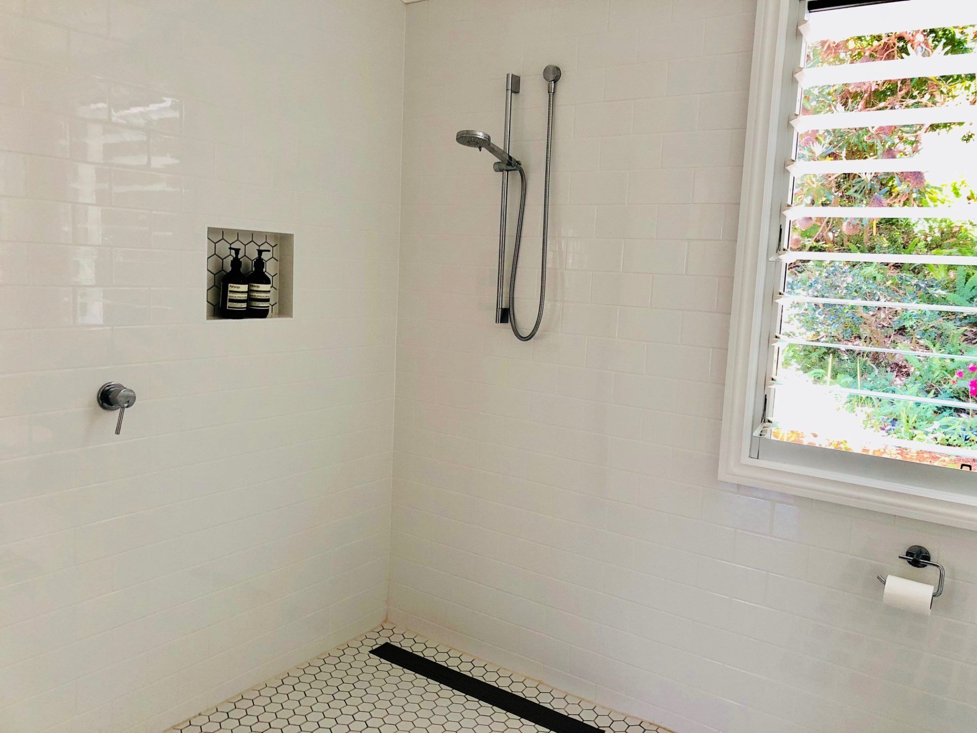 Ein Badezimmer mit weißen, sechseckigen Fliesen, einer Dusche inklusive Duschbrause, einem Toilettenpapierhalter und einem Fenster mit Jalousien.