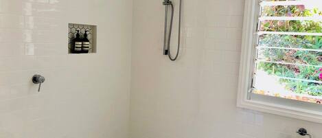 Ein Badezimmer mit weißen, sechseckigen Fliesen, einer Dusche inklusive Duschbrause, einem Toilettenpapierhalter und einem Fenster mit Jalousien.