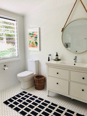 Une salle de bain avec des toilettes, un lavabo et un miroir. Il y a une fenêtre avec des stores, un panier en osier et un tapis noir et blanc sur le sol.