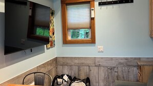 Ein kompakter Wohnraum mit Fernseher, einem Fenster mit Jalousien, einer Wandklimaanlage, einer Sitzecke und einem kleinen Tisch mit Fernbedienung.