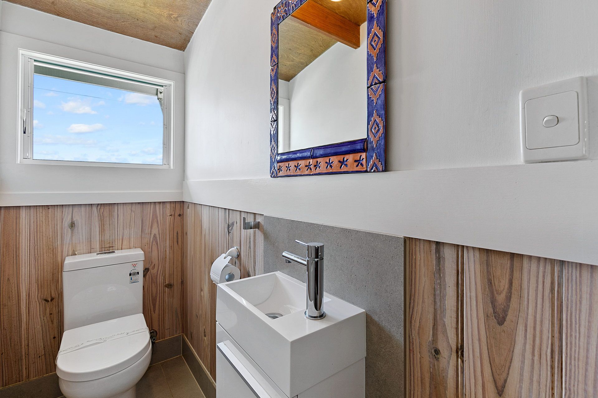 A bathroom with a wooden wall, a white toilet, a white sink with a chrome faucet, and a decorative mirror with a geometric pattern.