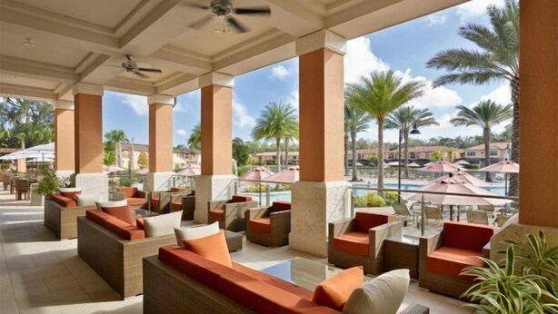 A covered outdoor seating area with wicker furniture and red cushions, overlooking a pool and palm trees.