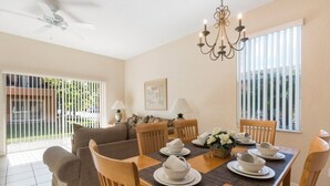 A dining area with a wooden table set for four, surrounded by chairs, a chandelier, and a view of the outdoors through large windows.