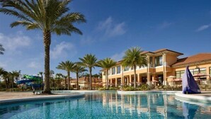 A swimming pool with palm trees, a water slide, and a hotel building with a red roof.