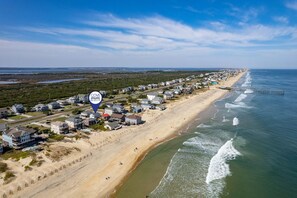 An aerial view of a coastal residential area with houses along the beach, a pier extending into the ocean, and a marker indicating a realty location.
