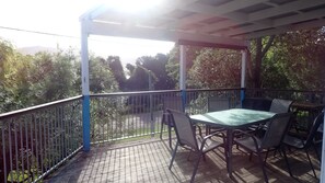 Une terrasse extérieure couverte, avec une table et des chaises, entourée d’une rambarde en métal et bordée d’arbres en arrière-plan.