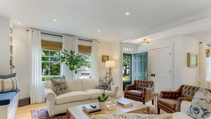 Un salon spacieux avec un canapé blanc, un fauteuil en cuir marron et une table basse. Il y a une grande fenêtre avec des rideaux, une plante et une porte avec un cadre bleu.