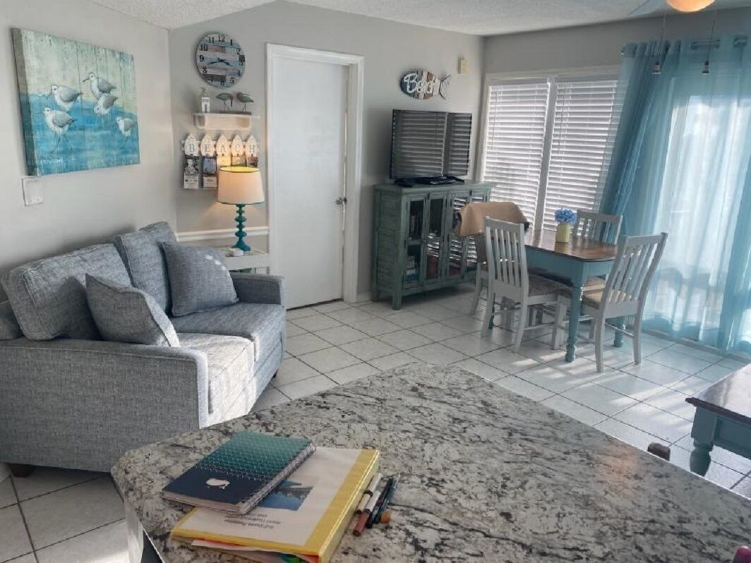 A living room with a grey sofa, a coffee table with books, a TV on a cabinet, and a dining area with chairs and a table.