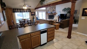 Una cocina con una isla central, armarios de madera y lavavajillas. Hay un comedor con sillas y una mesa, y una sala de estar con sofá y televisor.
