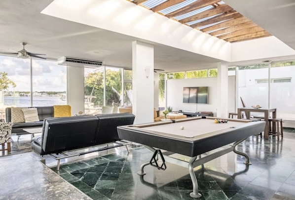 A spacious indoor area with a pool table, comfortable seating, and large windows offering views of the outdoors.