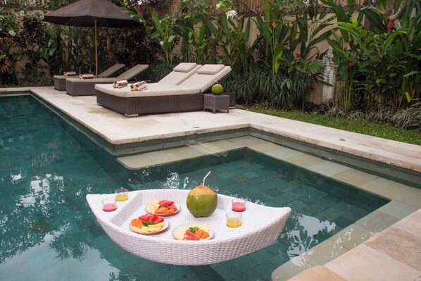 A poolside area with lounge chairs, a floating dining table with food and drinks, and lush greenery in the background. - Temple Estate; fully serviced 7 Bedroom villa, central Canggu close to the beach (Canggu)