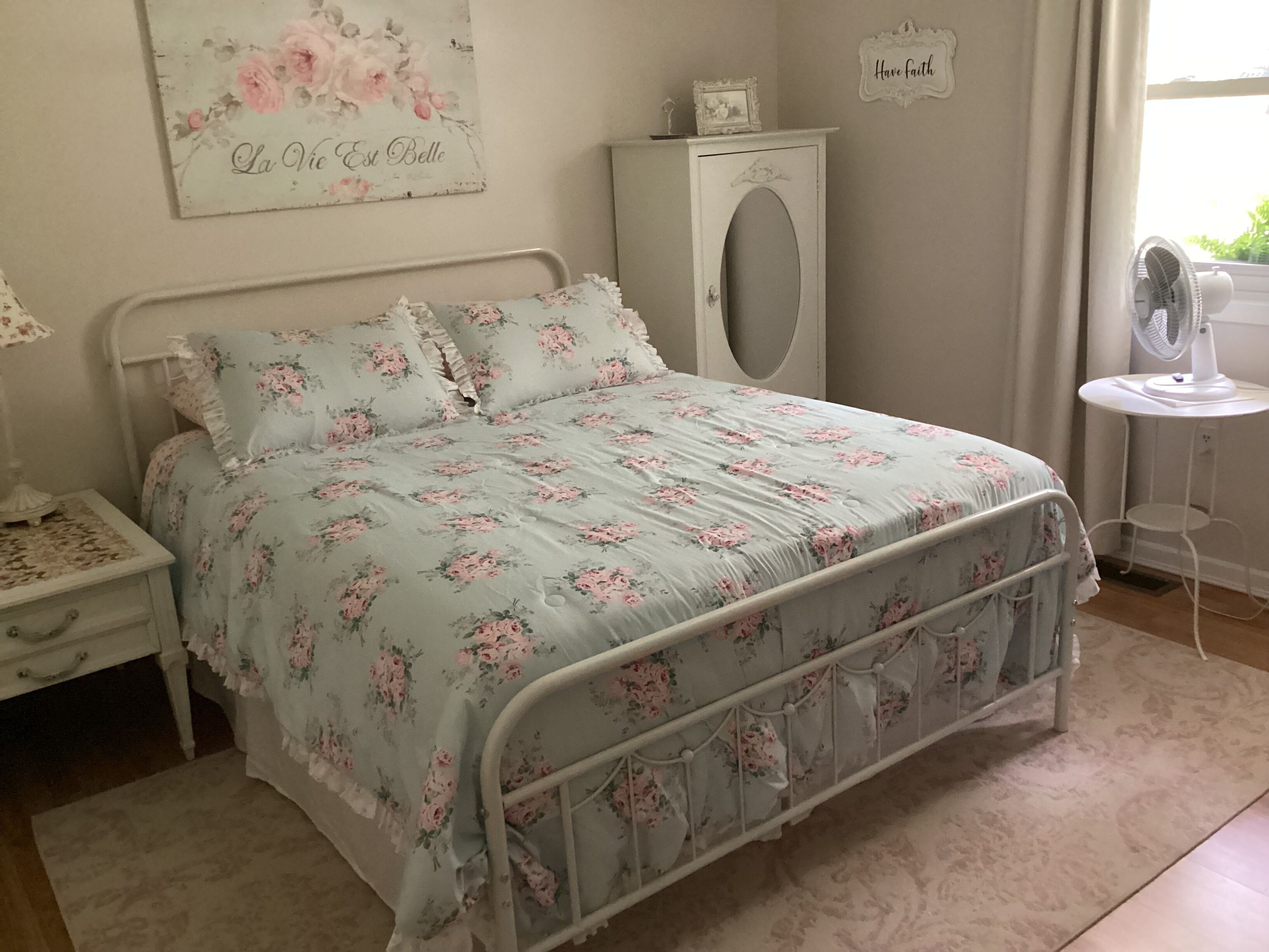 Quarto com cama estampada, mesa de cabeceira, cômoda, parede com letreiro floral e janela com vista.