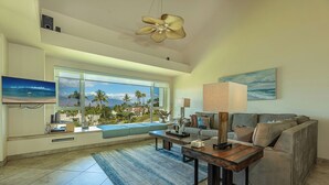 A spacious living room with a large window offering a view of palm trees and a beach. There is a sofa, a coffee table, and a television mounted on the wall. - Palms at Wailea  Spectacular Ocean Views with Exceptional Star Rating  (Kihei)