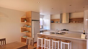 A modern kitchen with a central island, bar stools, wooden shelves, and a dining area with a table and chairs. - Rio Grande - views of the bay and the beach (CAMDEN HEAD)