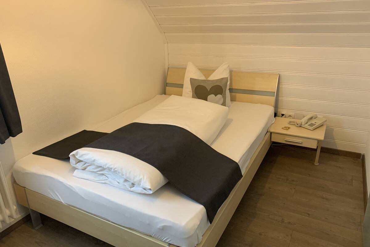Una cama individual con edredón blanco y negro, una mesita de noche con un teléfono, y suelo de madera.