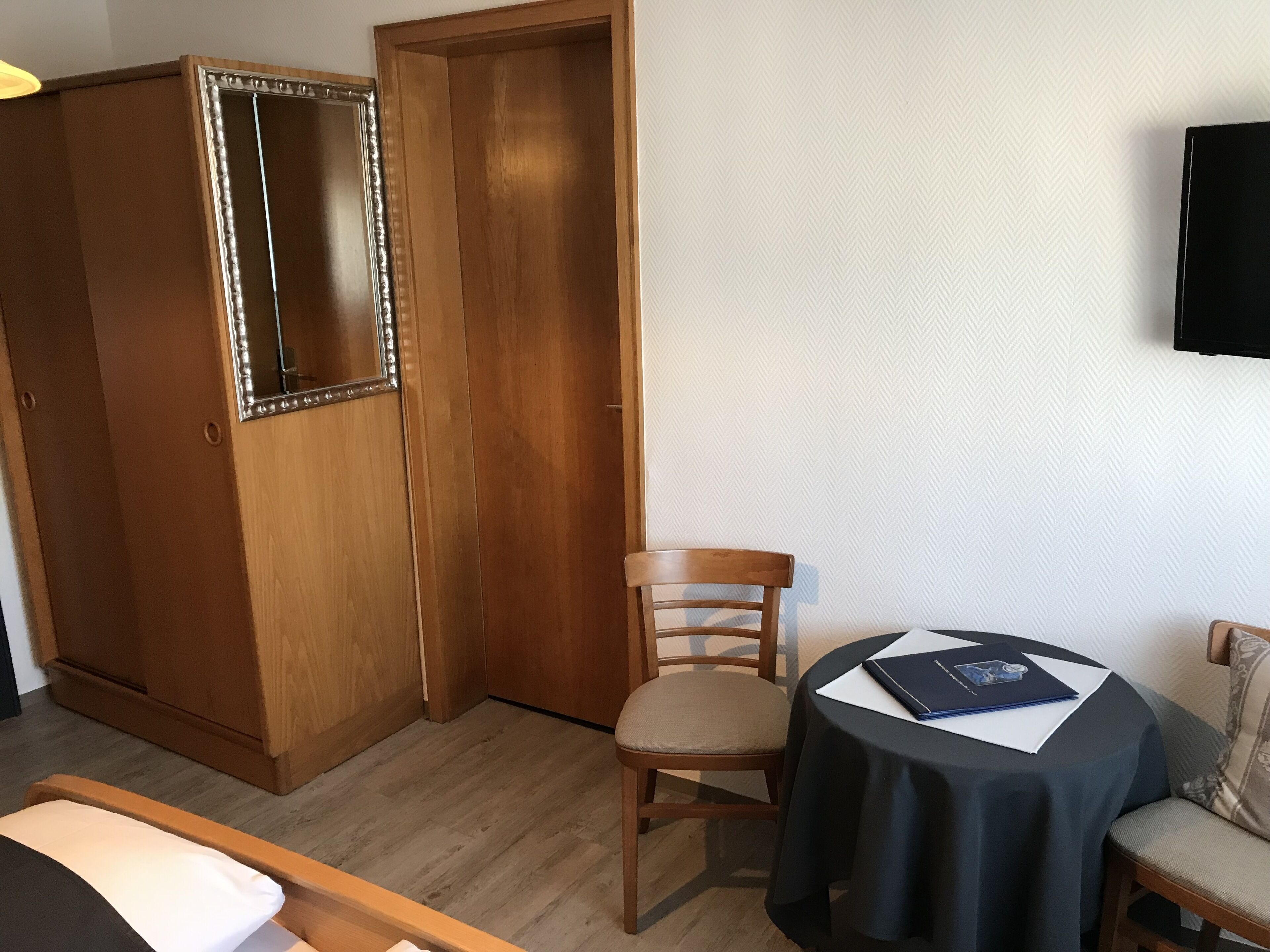 Una habitación de hotel con un armario de madera, una sola silla, una mesa redonda con mantel negro y un televisor montado en la pared.