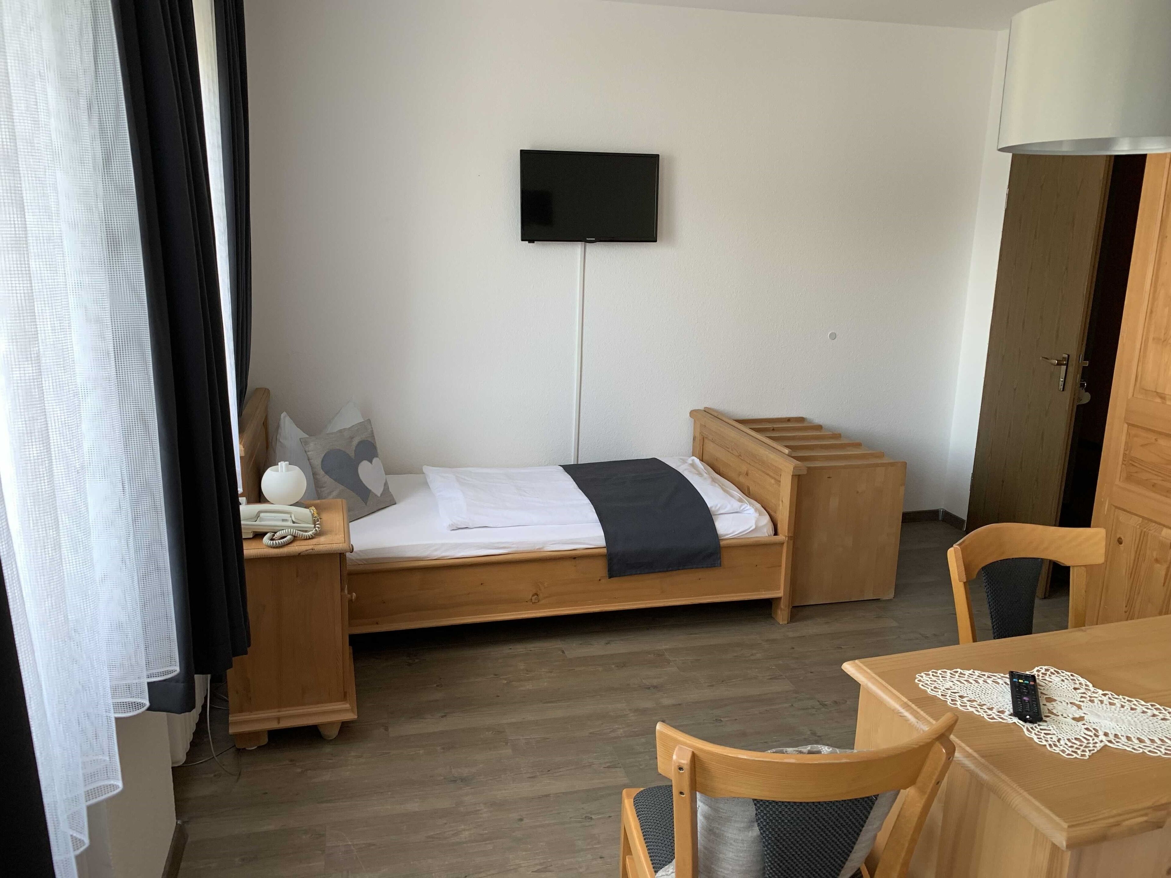 A single bed with a wooden headboard, a bedside table with a lamp, a TV mounted on the wall, and a wooden chair with a table.