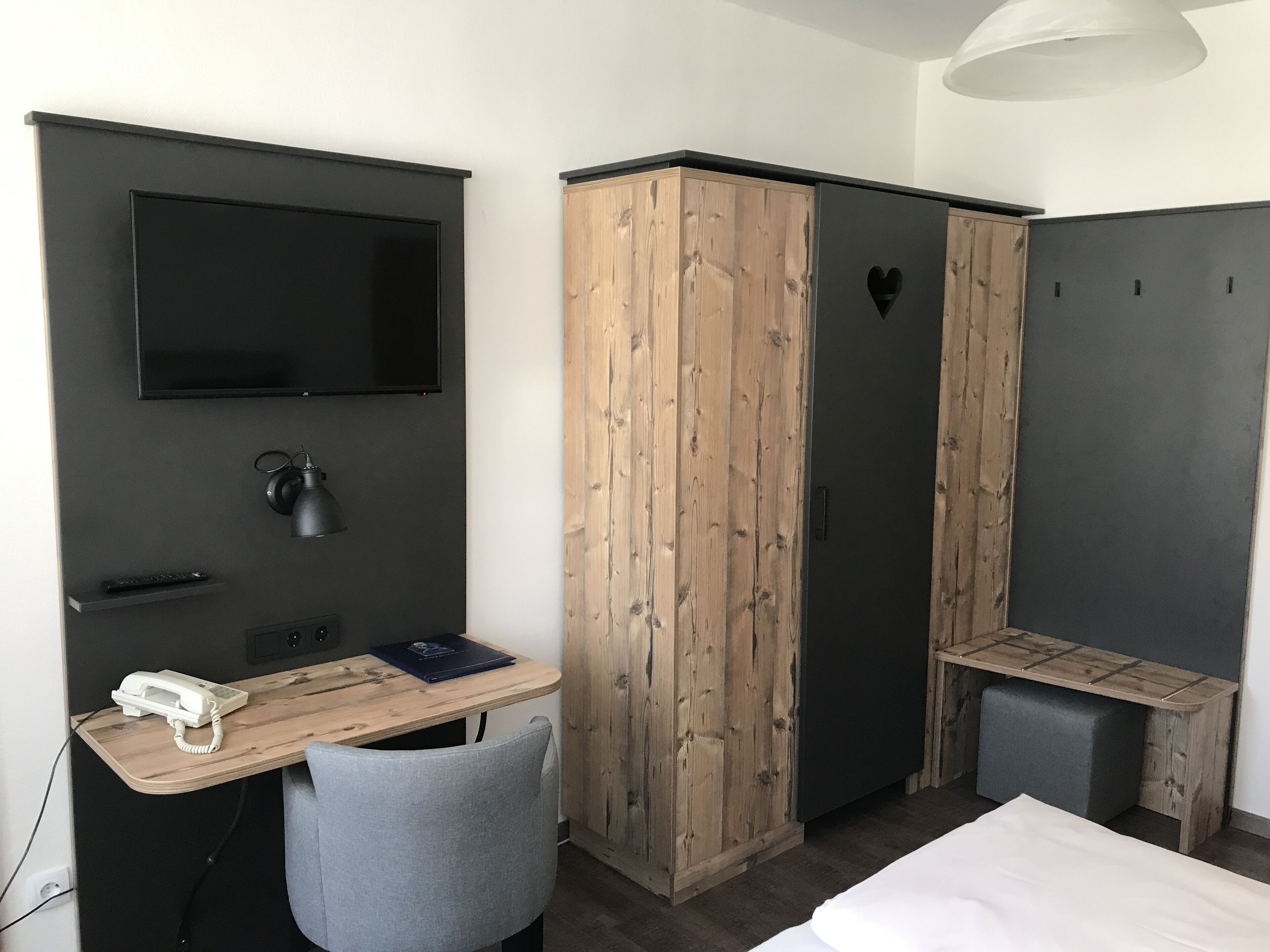 A compact hotel room with a bed, a desk with a chair, a television, a wardrobe, and a bedside table.