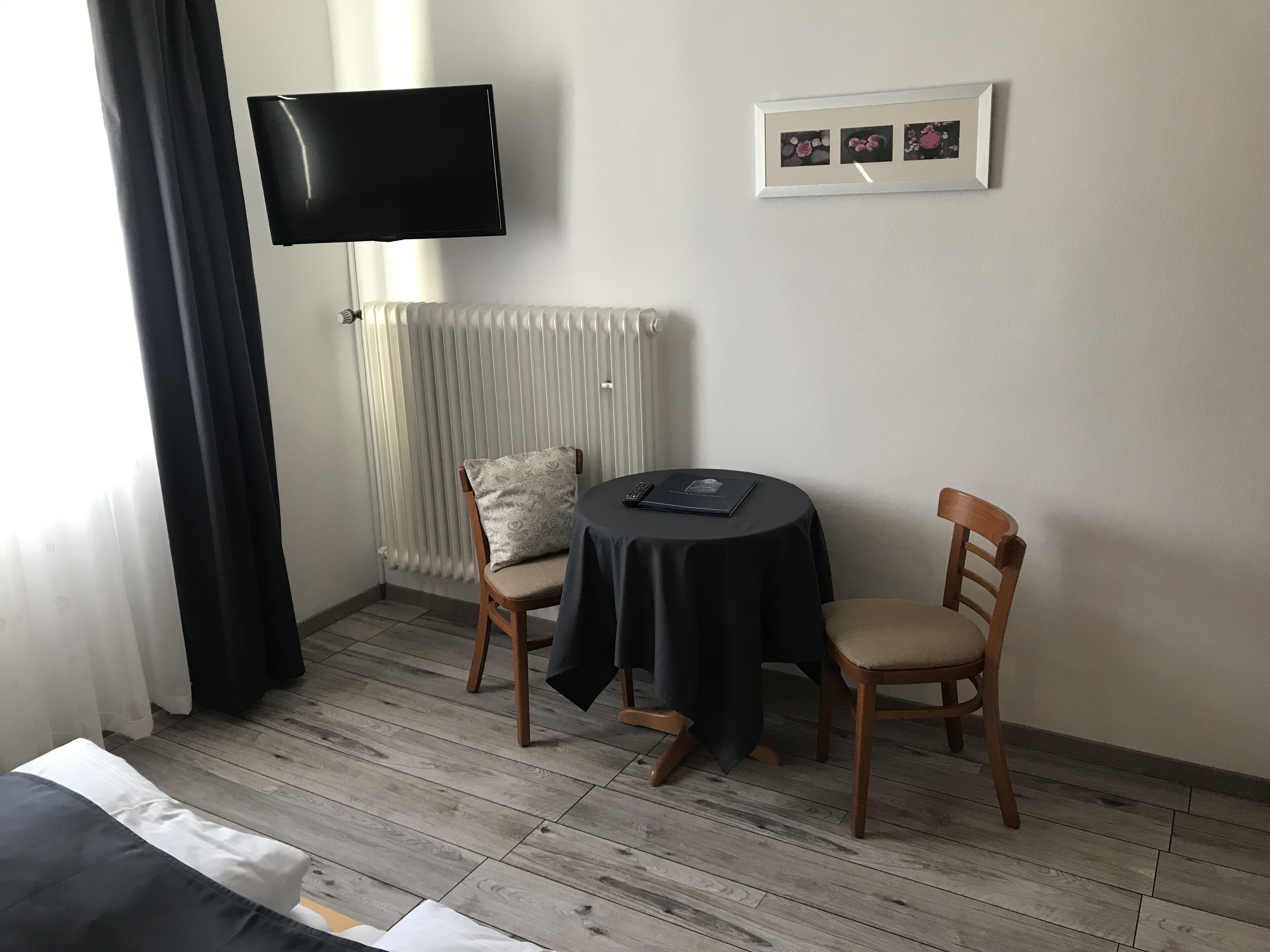 A hotel room with a bed, a TV mounted on the wall, a radiator, a small table with a chair, and a framed picture on the wall.