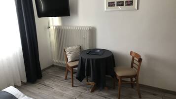A hotel room with a bed, a TV mounted on the wall, a radiator, a small table with a chair, and a framed picture on the wall.