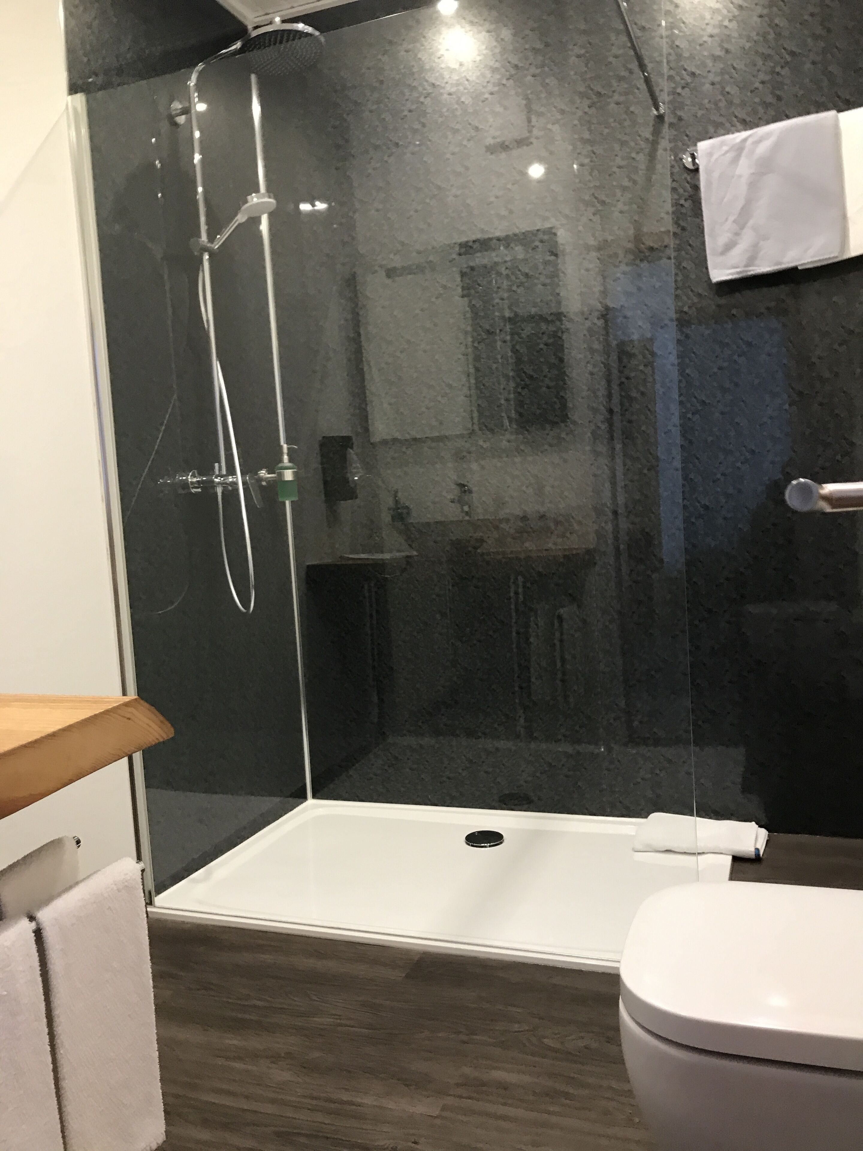 Un bany modern amb plat de dutxa envoltat de vidre, un vàter blanc i una prateleira de fusta.