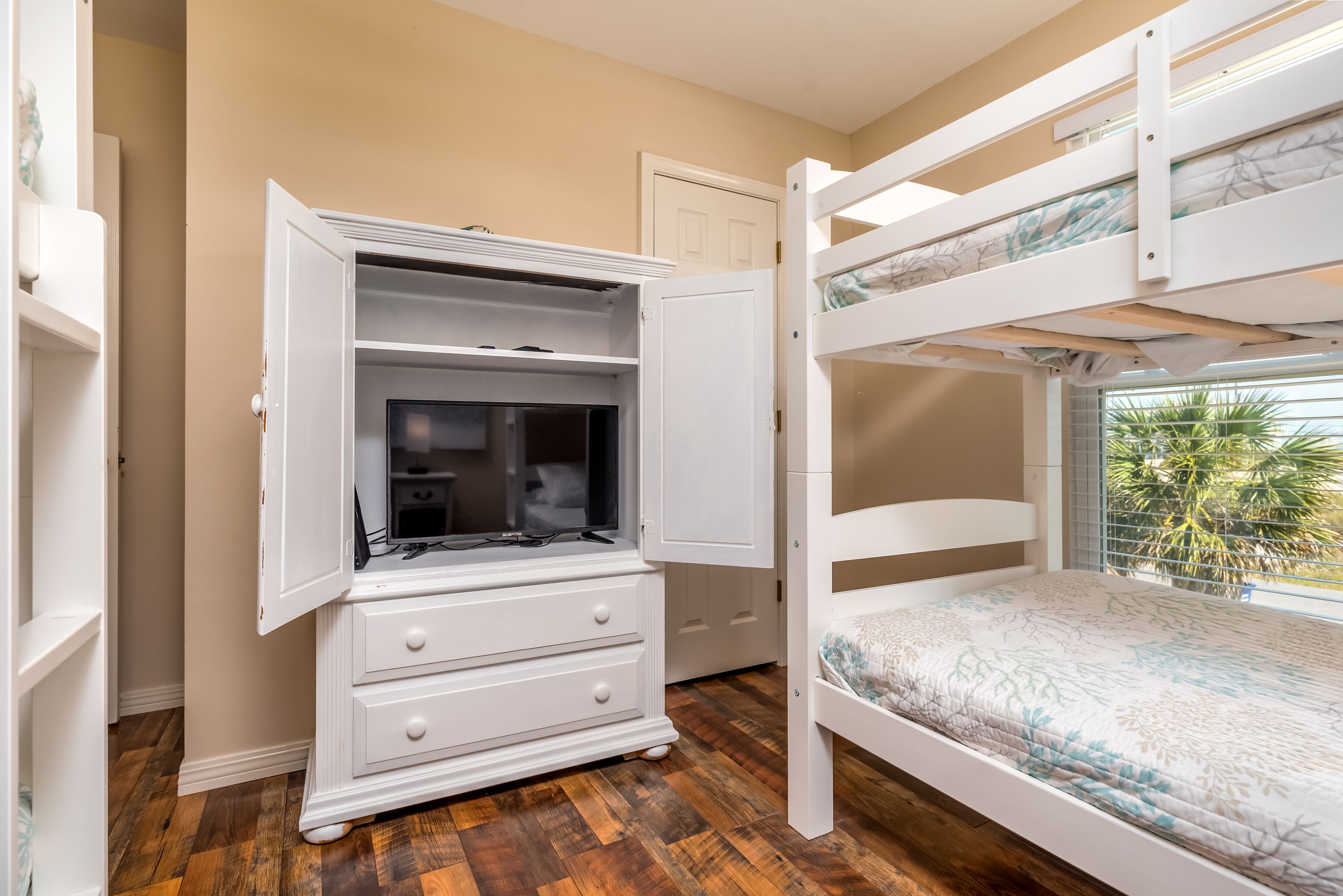 Una cama litera con ventana, un aparador blanco con cajones y una puerta en una habitación con suelo de madera.