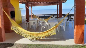 A covered outdoor seating area with white plastic chairs and tables, a yellow hammock, and a view of the sea. - Beachfront house with pool and Wi-Fi in Praia Azul - Pitimbu/PB (pitimbu)