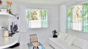 A modern living room with a white sofa, a glass coffee table, and a small dining table. There are two windows with bars, and a potted plant on the windowsill. - Beachfront house with pool and Wi-Fi in Praia Azul - Pitimbu/PB (pitimbu)