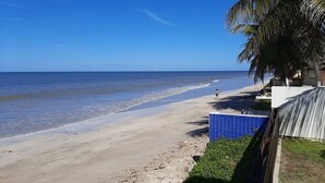 A beach with a clear sky, a sandy shore, and a few people walking. There is a blue fence and a palm tree visible. - Beachfront house with pool and Wi-Fi in Praia Azul - Pitimbu/PB (pitimbu)