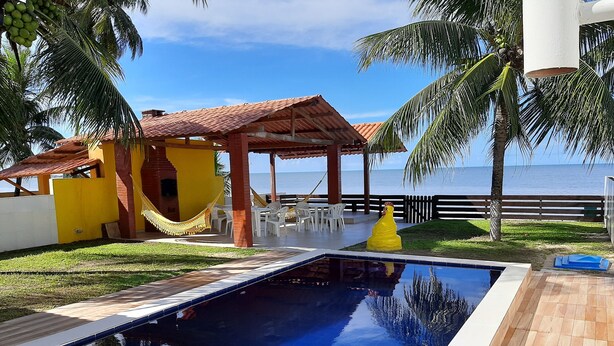 A pool area with a clear view of the sea, a yellow building, and a hammock. - Beachfront house with pool and Wi-Fi in Praia Azul - Pitimbu/PB (pitimbu)