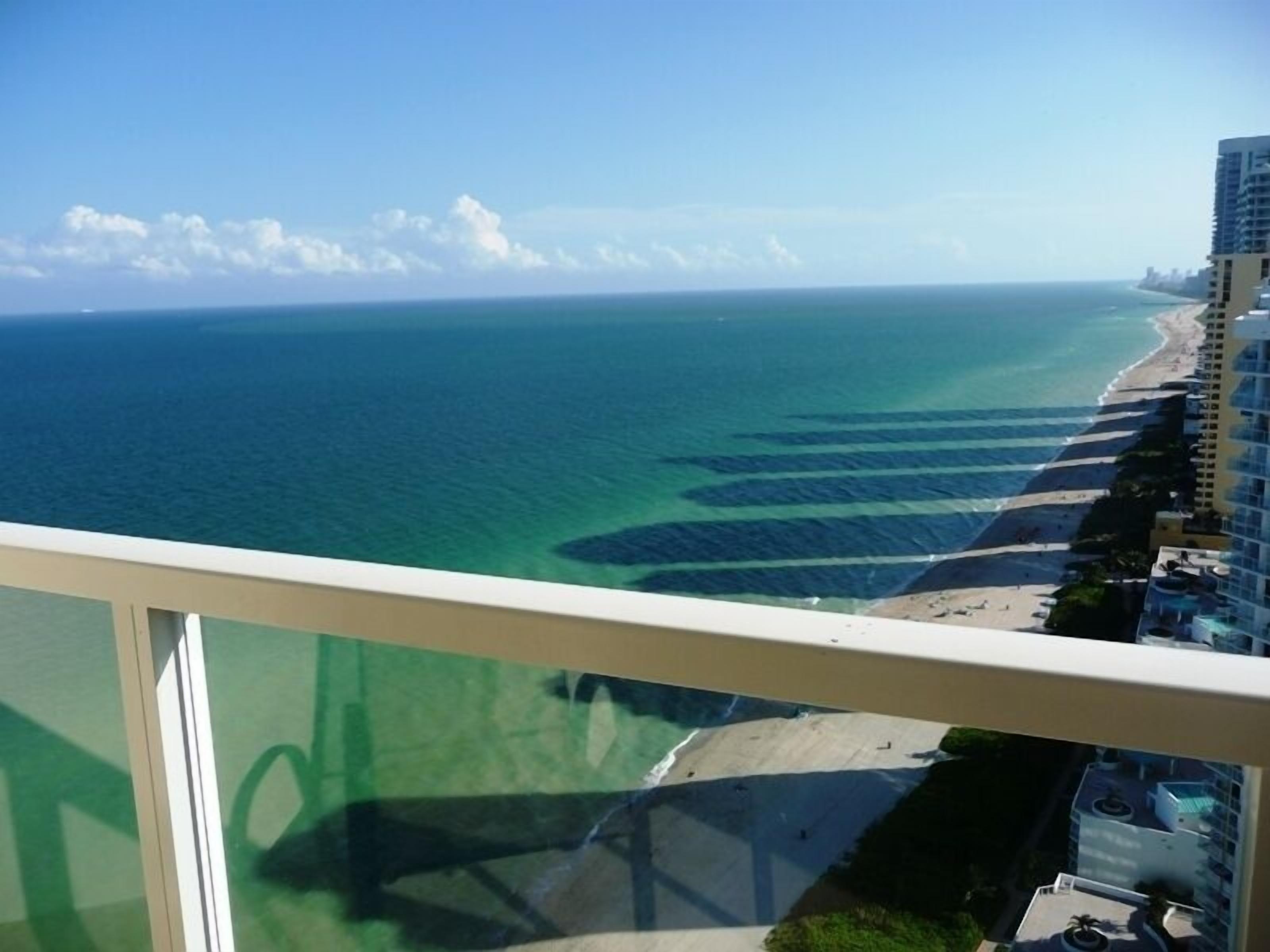 Une vue sur la côte, depuis un balcon donnant sur une plage aux eaux bleu azur, avec un hôtel situé à proximité du rivage.